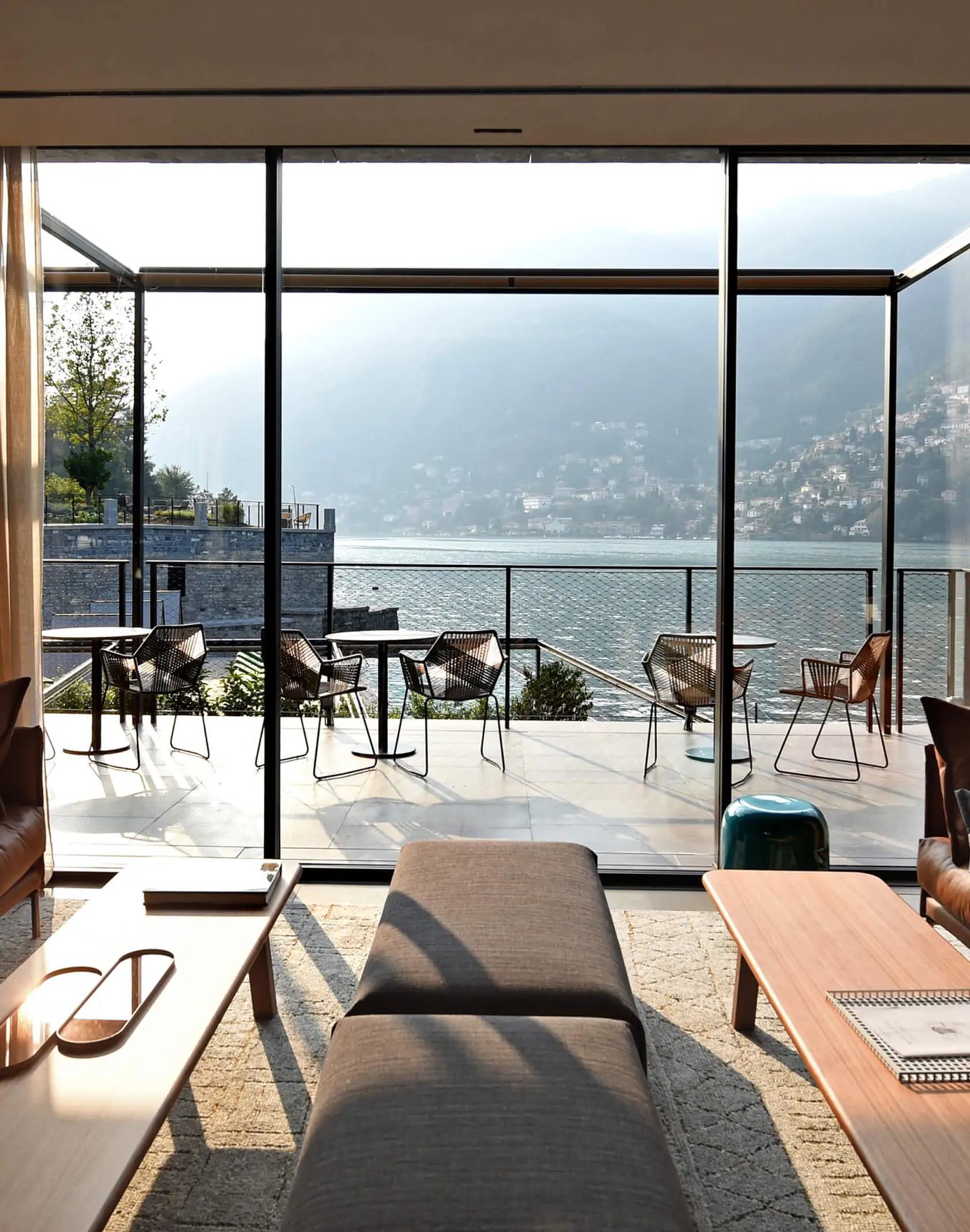 A light-filled lakeside lounge with floor-to-ceiling glass opening onto a terrace above the water — the kind of integrated hospitality environment where real estate, design and experience converge.